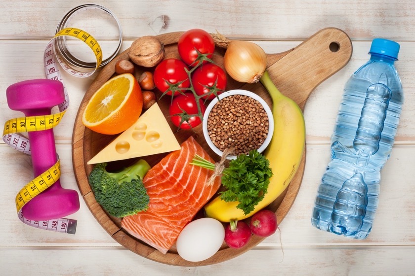 Holzbrett mit Lachs, Gemüse, Obst, Käse und Buchweizen, daneben Hantel, Maßband und Wasserflasche